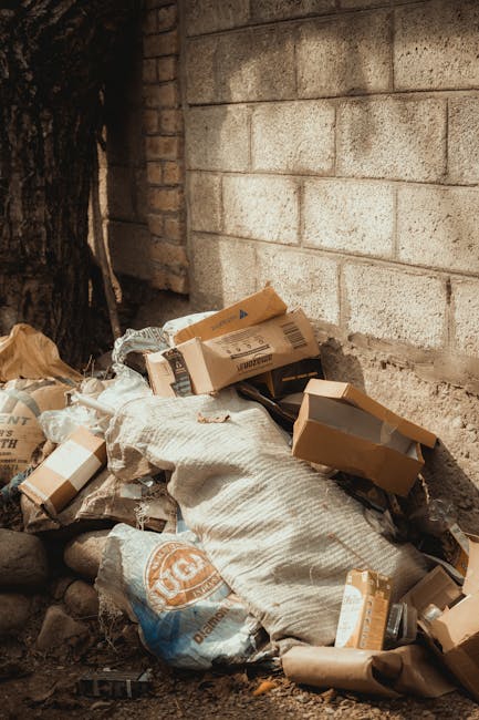 A pile of mixed waste materials is situated at the base of a brick wall, adjacent to a rough tree trunk on the left side of the image. The waste includes several cardboard boxes, some partially collapsed or open, with visible tape and printed labels, along with white and beige fabric or shredded packaging material beneath the boxes. Surrounding the debris are small stones and dirt, indicating an outdoor setting, possibly a back alley or yard. The brick wall in the background is constructed of large, beige-colored concrete blocks with a rough texture and some slight staining or weathering. To the left, part of a dark tree trunk is visible, providing natural contrast to the man-made structures. The scene is lit by natural daylight, highlighting the textures of the cardboard, fabric, and brick, conveying an environment related to waste disposal or rubbish accumulation, which may be targeted for removal by services like Rubbish Removal Barnes, supporting private waste management or alternative rubbish collection options.