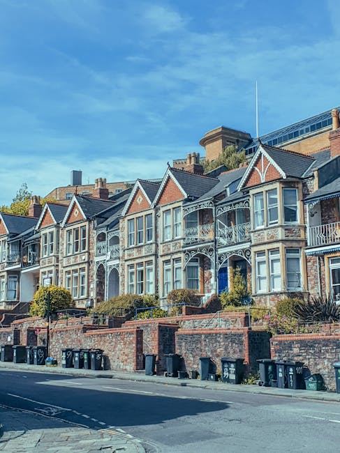 A row of Victorian-style residential houses situated on a sloped street with brick facades, decorative white trim, bay windows, and ornate ironwork balconies. The houses are set behind a low brick wall with a paved sidewalk in front. Several waste bins are lined up along the curb, indicating trash collection or waste management activity typical of private rubbish removal services. The background shows a partly cloudy sky with blue tones, and some modern buildings can be seen behind the row of houses. The scene exemplifies urban residential architecture with a focus on exterior details, highlighting the context of local rubbish disposal options in private properties, supporting independent waste collection or on-site clearance activities.