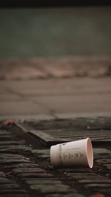 A disposable paper cup with a white exterior and printed text lies abandoned on a weathered brick pavement outdoors, near a metal grate or drain cover. The cup appears to be empty and is resting on its side, with a slight tilt. The surrounding environment includes a concrete or stone surface and a background of blurred greenish-gray tones, suggesting an outdoor setting such as a street or alleyway. The diffuse lighting indicates it may be an overcast day. This scene relates to waste disposal practices and highlights the importance of proper rubbish removal, potentially handled by services like Rubbish Removal Barnes, which supports efficient and environmentally responsible waste collection in residential or commercial areas.