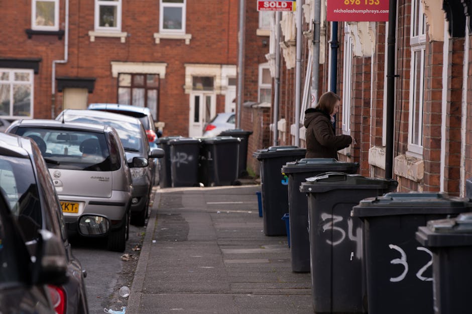 A narrow residential street in Barnes shows multiple black wheelie bins lined up along the sidewalk in front of red-brick terraced houses. The bins are made of plastic with textured surfaces, some with lifted lids, and are numbered with white spray paint. A woman dressed in a dark coat stands near the bins, appearing to be opening a lid or preparing to dispose of waste. To the left, parked cars occupy the curb with visible rear windows, taillights, and license plates. The pavement is a typical grey concrete slab, and the surrounding environment includes brick facades with white window frames and decorative stone lintels above some windows. In the background, additional cars and more houses can be seen, with natural daylight illuminating the scene and casting soft shadows. This setup reflects typical private waste disposal arrangements outside residential properties, consistent with services by Rubbish Removal Barnes specializing in independent rubbish collection and on-site clearance.